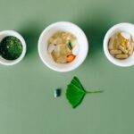 tisso - Flatlay of capsules and herbs in ceramic bowls on green surface.